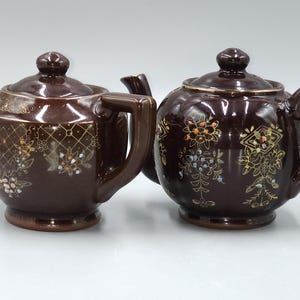 May include: Two dark brown ceramic teapots with matching lids. The left teapot features a gold grid pattern and floral accents. The right teapot has a ribbed body with gold floral designs. Both have curved spouts, handles, and circular bases.