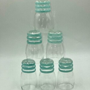 May include: A set of six clear glass tumblers arranged in a pyramid formation. Each tumbler features a rounded body and a neck with three distinct horizontal teal-colored rings encircling the top rim, providing a consistent decorative accent.