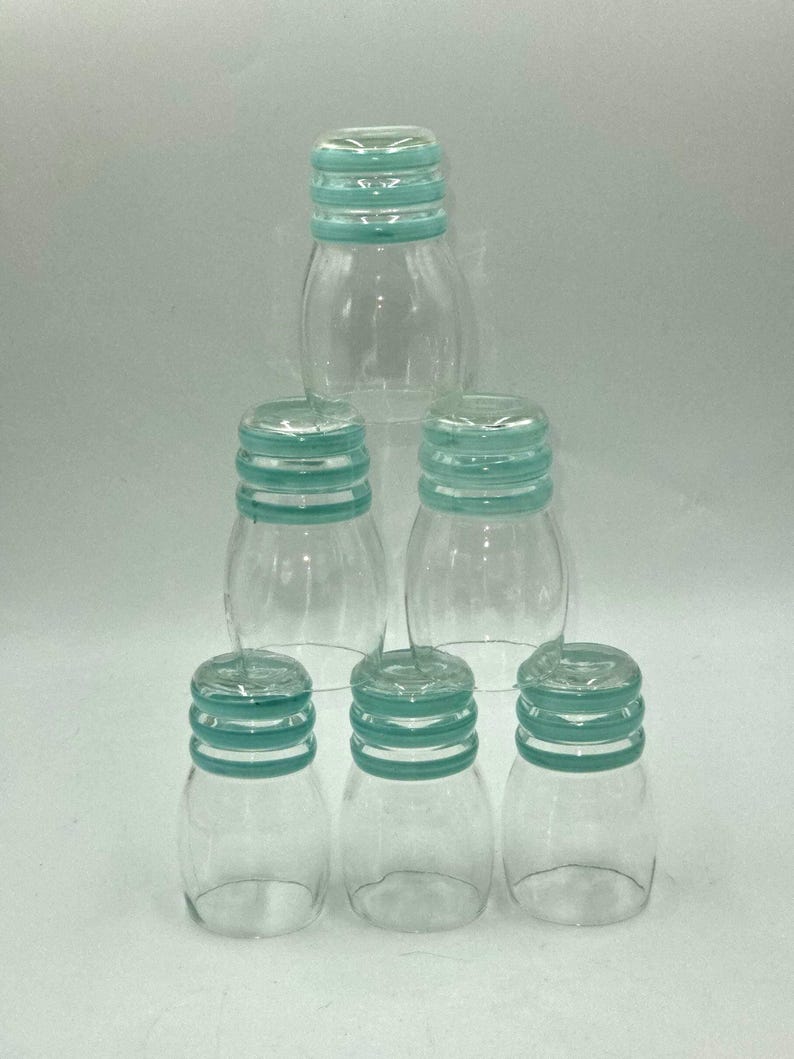 May include: A set of six clear glass tumblers arranged in a pyramid formation. Each tumbler features a rounded body and a neck with three distinct horizontal teal-colored rings encircling the top rim, providing a consistent decorative accent.