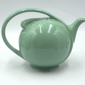 Puede incluir: Una tetera de cerámica verde menta con un asa curva y un pico. La tetera tiene un cuerpo redondeado y una tapa con un pequeño asa. La tetera está sobre un fondo blanco.