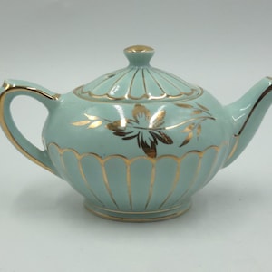 May include: A light blue teapot featuring a gold-trimmed scalloped pattern along the base and lid. The body is decorated with a gold floral motif on the side, and the handle, spout, and lid finial are accented with gold trim.