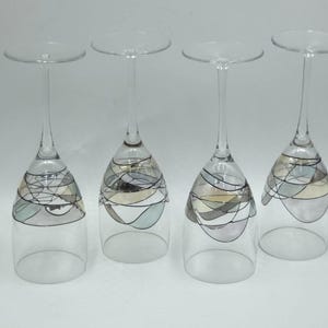 May include: Four clear glass wine glasses standing upside down. Each glass features a hand-painted design on the bowl consisting of thin black lines forming abstract, overlapping geometric shapes with muted blue, gray, and beige color fills.