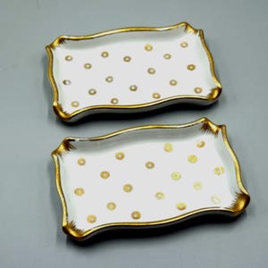 Pode incluir: Duas bandejas retangulares de porcelana branca com bordas douradas e bolinhas douradas. As bandejas têm um design de borda ondulada. As bandejas são provavelmente para servir ou exibir.
