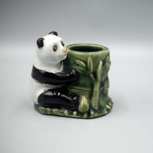 Könnte beinhalten: Eine Keramik-Panda-Figur, die einen grünen Bambus-Pflanztopf hält. Der Panda ist schwarz-weiß mit einem grünen Sockel.