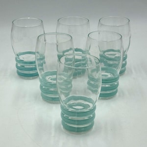 May include: A set of six clear glass tumblers arranged in a triangular formation. Each tumbler features a rounded body and a base decorated with three horizontal, raised teal-colored rings that provide a textured grip around the lower section.