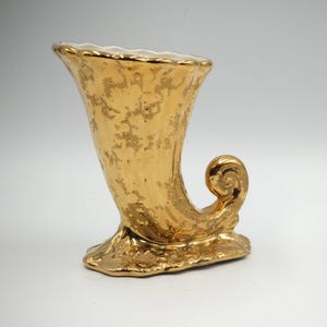 Può includere: Un vaso in ceramica dorata a forma di corno con un manico ricurvo. Il vaso ha una superficie testurizzata e un interno bianco.