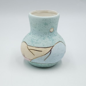 Puede incluir: Un pequeño jarrón de cerámica azul claro con textura moteada. El jarrón presenta un cuerpo redondeado, un cuello estrecho y un interior blanco. Tiene un diseño decorativo en beige, azul claro y detalles dorados.