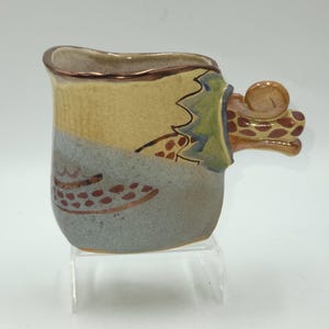 May include: A ceramic mug with a unique design. The mug features a tan, blue, and green colour scheme with a decorative handle. The handle has a giraffe-like pattern. The mug is approximately 10 cm tall.
