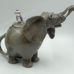 Puede incluir: Una tetera de cerámica gris brillante con forma de elefante, trompa levantada y colmillos blancos. Una pequeña figura de pájaro morado y blanco se asienta sobre la tapa, que forma el lomo del elefante. La tetera tiene un asa curva y cuatro patas.