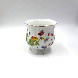 May include: White ceramic planter with a scalloped rim and gold trim. The planter is decorated with colorful illustrations of strawberries, butterflies, and insects. The planter is approximately 4 inches tall.