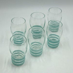 May include: A set of five clear glass tumblers, each featuring a rounded body and a base decorated with three horizontal, raised light blue rings. The glasses have a smooth, transparent upper section and a textured, ribbed lower portion.