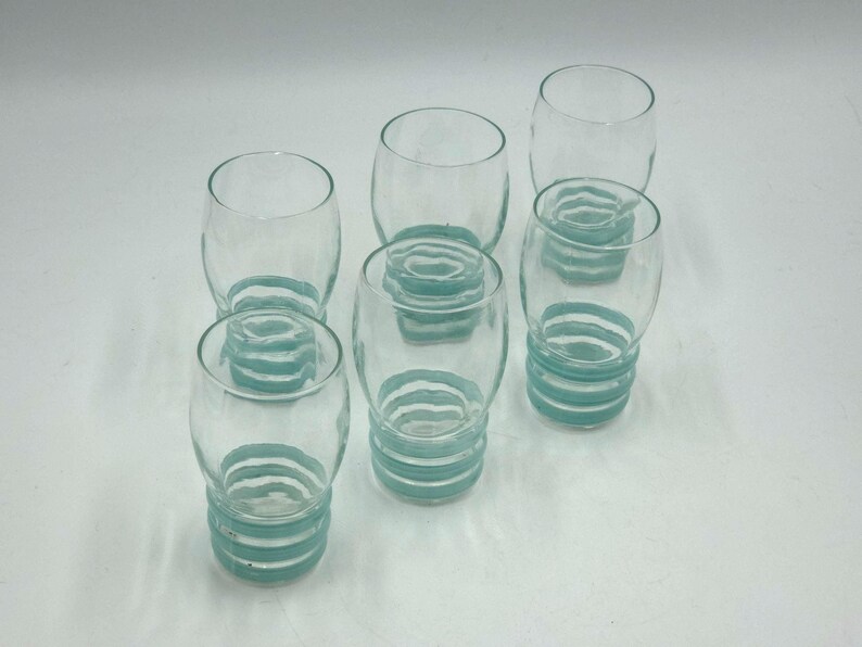 May include: A set of five clear glass tumblers, each featuring a rounded body and a base decorated with three horizontal, raised light blue rings. The glasses have a smooth, transparent upper section and a textured, ribbed lower portion.
