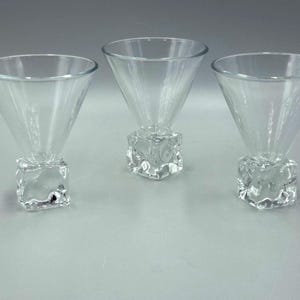 May include: Three clear cocktail glasses. Each glass has a conical bowl and a square, textured base. The glasses are arranged in a row, highlighting their design and clarity. Suitable for serving cocktails.