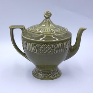 May include: Olive green ceramic teapot with a lid and handle. The teapot has a decorative pattern around the body and on the lid. The spout is curved, and the handle is a simple loop. The teapot sits on a small pedestal base.