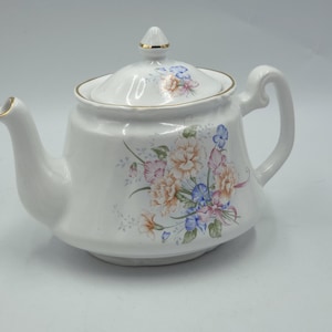 Könnte beinhalten: Weiße Keramik-Teekanne mit Deckel, Henkel und Ausguss. Die Teekanne ist mit einem Blumenmuster in Rosa-, Blau- und Orangetönen verziert. Goldfarbene Verzierungen akzentuieren den Rand, den Ausguss und den Deckel. Ein klassisches Teezubehör.