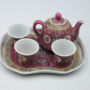 May include: A pink and white ceramic tea set comprising a teapot, three cups, and a serving tray. The set features a floral and geometric design with Chinese characters. The teapot has a curved handle and spout. The tray has a scalloped edge.