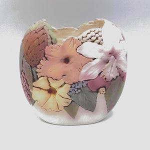 May include: A ceramic vase with a floral design. The vase features a variety of flowers in shades of pink, peach, and yellow, with green leaves and a textured surface. The vase has an oval shape and is set against a white background.