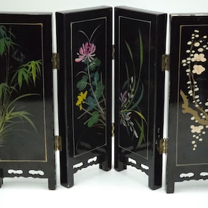 May include: A four-panel black lacquer folding screen featuring hand-painted botanical illustrations. Each panel displays different flora, including green bamboo, pink and yellow flowers, tall grasses, and white blossoms on a brown branch.