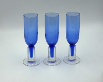 May include: Three blue glass champagne flutes. Each flute has a tall, cylindrical bowl and a short stem with a round base. The glass has a gradient effect, transitioning from a darker blue at the top to a lighter blue at the base.