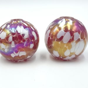 May include: Two spherical glass beads featuring an iridescent finish with a mottled pattern of white, gold, and magenta patches. Each bead has a small circular opening at the top, showcasing a smooth, reflective surface and a multi-colored design.
