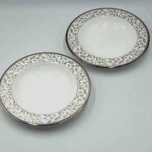 Könnte beinhalten: Zwei weiße Keramikschalen mit silberfarbenem Rand und einem dekorativen grauen Blumenmuster am Rand. Die Schalen sind rund und haben eine geringe Tiefe, geeignet zum Servieren von Suppen oder Salaten.