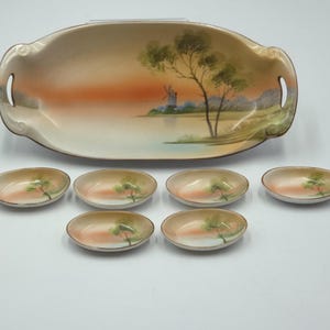 May include: A vintage porcelain serving set featuring a large oval tray with handles and six smaller oval dishes. The set is painted with a landscape scene of a windmill, trees, and a sunset in shades of orange, green, and beige.
