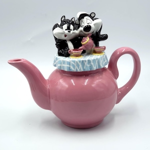 Puede incluir: Una tetera de cerámica rosa con una tapa rematada por dos figuras de mofetas en blanco y negro sentadas a una mesa con un mantel de cuadros azules y blancos. Las mofetas aparecen con una pequeña tetera rosa y dos tazas de té a juego.