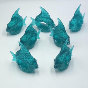 Puede incluir: Colección de ocho figuras de peces de vidrio turquesa. Cada pez tiene aletas detalladas y un cuerpo texturizado. Las figuras están dispuestas sobre una superficie blanca, mostrando su diseño acuático.