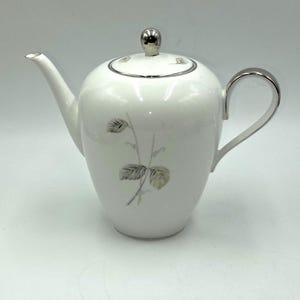 May include: White porcelain teapot with silver accents, including the handle, spout, and lid. The teapot features a delicate floral design with gray and gold leaves. A classic piece for serving tea.