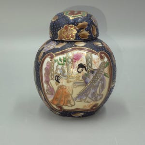 May include: A round, lidded ceramic jar with a dark blue background and gold floral accents. The jar features a painted scene of figures in a room, with details in orange, purple, and white. The lid is also dark blue with gold details.