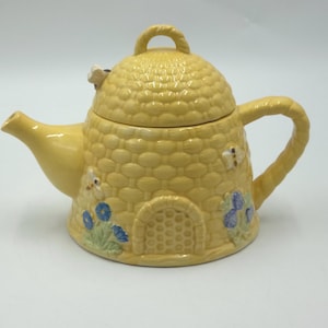 May include: Yellow ceramic teapot shaped like a beehive, with a matching lid and handle. The teapot is decorated with small butterflies and blue flowers. The spout and handle are curved. The teapot is a charming addition to any kitchen.