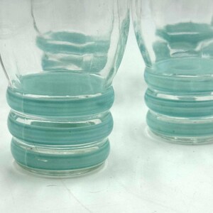 May include: Two clear glass tumblers featuring three horizontal, raised teal-colored rings around the lower portion of each vessel. The glass has a smooth, transparent finish above the textured, colored bands at the base of each drinking glass.