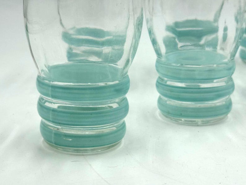 May include: Two clear glass tumblers featuring three horizontal, raised teal-colored rings around the lower portion of each vessel. The glass has a smooth, transparent finish above the textured, colored bands at the base of each drinking glass.