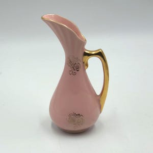 Puede incluir: Una jarra de cerámica rosa con asa y borde dorados. La jarra tiene detalles florales dorados y un pico curvo. El asa es dorada y el interior del pico también es dorado.