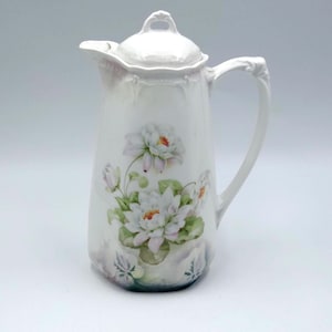 Puede incluir: Jarra de porcelana blanca con tapa, con un diseño floral de nenúfares blancos con centros naranjas y hojas verdes. La jarra tiene un asa curva y un pico vertedor. Una pieza decorativa para servir bebidas.