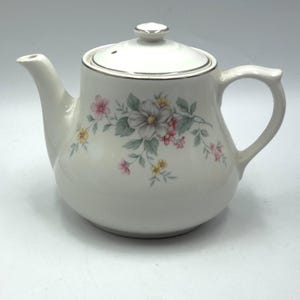 Könnte beinhalten: Weiße Keramik-Teekanne mit Deckel und Henkel. Die Teekanne ist mit einem Blumenmuster in Rosa, Gelb und Grau verziert. Der Rand der Teekanne und des Deckels ist silberfarben.