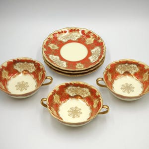 Może przedstawiać: Zestaw czterech porcelanowych misek do zupy z pozłacanymi brzegami i misternymi czerwonymi i złotymi wzorami kwiatowymi. Miski mają dwa uchwyty i idealnie nadają się do serwowania zupy lub deseru.