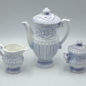 May include: A three-piece ceramic tea set in a light blue and white color scheme. The set includes a teapot with a lid, a creamer, and a sugar bowl with a lid. Each piece features ornate embossed floral and scrollwork detailing.