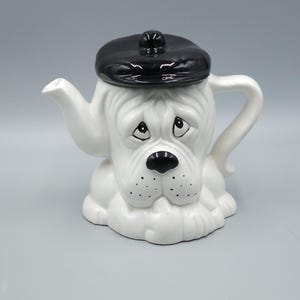 Puede incluir: Una tetera de cerámica blanca con forma de cabeza de perro, con una boina negra. La tetera tiene un pico, un asa y una nariz negra. La cara del perro tiene una expresión triste con ojos y puntos negros.