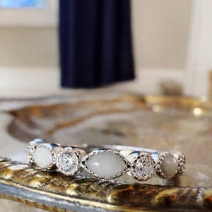 May include: A silver ring featuring a series of alternating gemstones and clear, round-cut stones. The gemstones are oval-shaped and have a milky white appearance. The ring is set on a decorative, gold-toned surface.