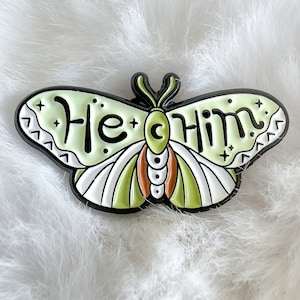 Puede incluir: Pin de esmalte verde y blanco con forma de mariposa con las palabras "He/Him" en las alas.