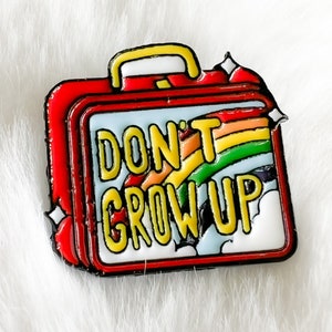Könnte beinhalten: Rote, gelbe und blaue Emaille-Anstecknadel in Form einer Vintage-Lunchbox mit einem Regenbogen und dem Text "DON'T GROW UP".