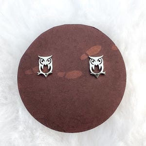 May include: A pair of silver owl stud earrings on a brown background.