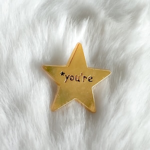 Puede incluir: Un amuleto dorado en forma de estrella con la palabra "you're" inscrita en él.