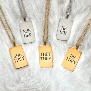 May include: Five gold and silver necklaces with rectangular pendants. The pendants have the words "She Her", "They Them", "He Him", "She They", and "He They" written on them.