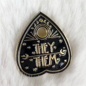 Peut inclure: Épinglette en forme de cœur noir et or avec les mots "They Them" en lettres dorées. L'épingle a une lune croissante et des étoiles autour des mots.
