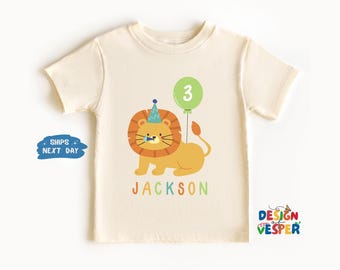 May include: Cream-colored t-shirt featuring a cartoon lion wearing a party hat and holding a green balloon with the number 3. The name "JACKSON" is printed in colorful letters below the lion. A fun birthday shirt.