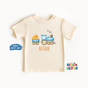 May include: Cream-colored t-shirt featuring a watercolor illustration of a blue train pulling a cart of pumpkins. The name "ARTHUR" is printed in orange below the train. The shirt has short sleeves and a crew neck.
