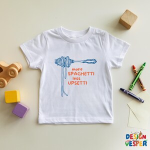 Pu&ograve; includere: Maglietta bianca con grafica blu di spaghetti e forchetta e la scritta arancione "more spaghetti less upsetti". L'immagine include giocattoli e pastelli per bambini.