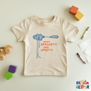 Pu&ograve; includere: Una maglietta beige con la scritta "more spaghetti less upsetti" in blu e arancione. La grafica mostra una forchetta con spaghetti. La maglietta &egrave; circondata da giocattoli e pastelli colorati.